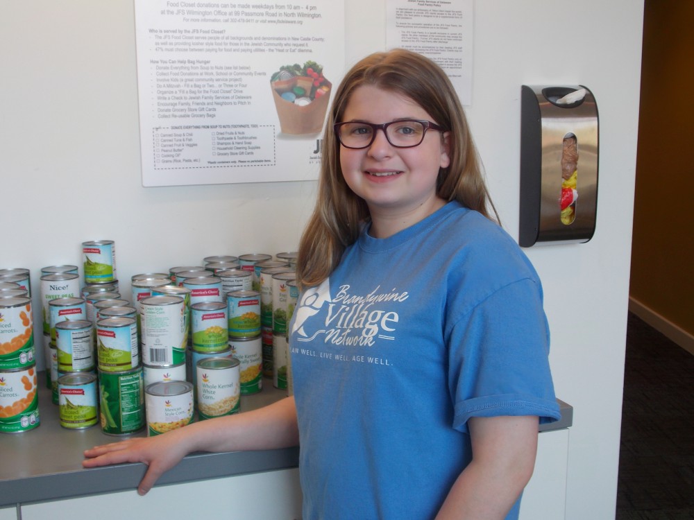 A young volunteer in a light blue shirt stands in front of the JFS Food Pantry, where she has stocked canned goods and toiletries.